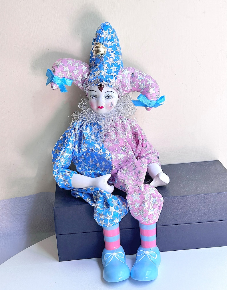 May include: A porcelain jester doll with a blue and pink star patterned costume, a blue and white striped hat with a bell, and blue shoes with white bows.