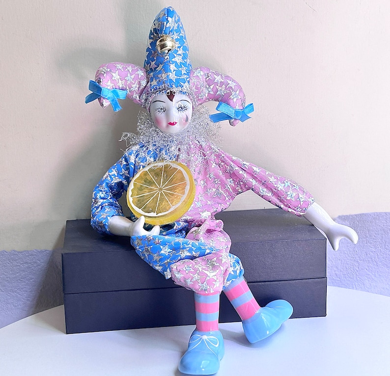 May include: A porcelain doll dressed as a jester with a blue and pink star patterned costume, holding a yellow lemon slice. The doll is sitting on a black box.