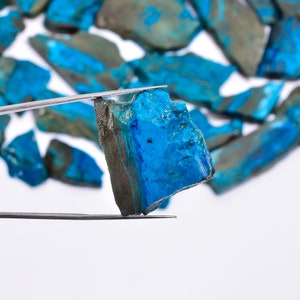 May include: A collection of raw, natural chrysocolla stones in shades of blue and green. One stone is held by tweezers, showcasing its vibrant color and rough texture. The background features more of the same stones.
