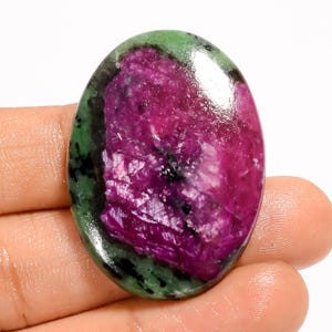 May include: Oval-shaped gemstone with a vibrant mix of colors. The stone features a deep magenta center, surrounded by a band of green and black. The polished surface reflects light, highlighting the natural patterns and textures of the stone.