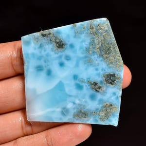 Puede incluir: Una pieza pulida de piedra preciosa de larimar de forma irregular. La piedra presenta un color azul claro con vetas blancas y parches de inclusiones grises y marrones. La piedra preciosa se sostiene en una mano sobre un fondo negro.