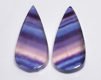 May include: Two teardrop-shaped fluorite gemstones. The stones display a gradient of colors, including purple, blue, and white, with horizontal banding. The polished stones are set against a white background.