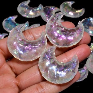 Puede incluir: Una colección de cristales blancos iridiscentes en forma de media luna. Los cristales tienen un brillo arcoíris y están dispuestos en una mano.
