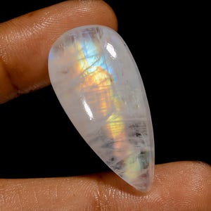 May include: A pear-shaped, polished moonstone gemstone. The stone is primarily white with iridescent flashes of blue, yellow, and orange. The gemstone is held between fingers against a black background.