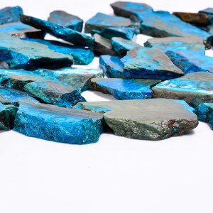 May include: A collection of raw, irregularly shaped stones with a vibrant blue hue, interspersed with areas of teal and gray. The stones are scattered against a white background, showcasing their natural textures and colors. These stones could be used for jewelry making or decorative purposes.