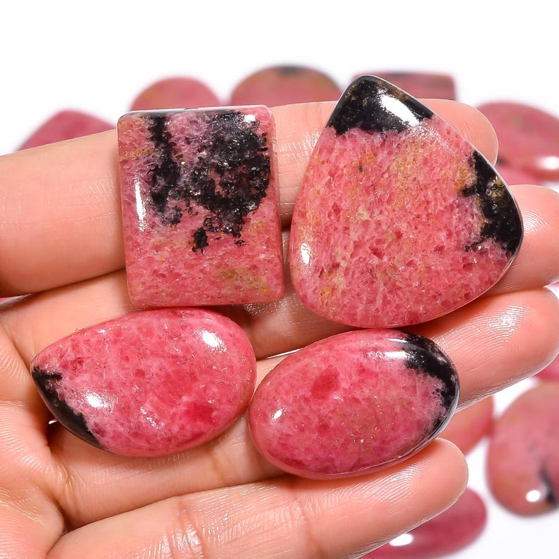 May include: Assortment of pink and black gemstone cabochons. The cabochons are cut in various shapes, including oval, teardrop, and square.