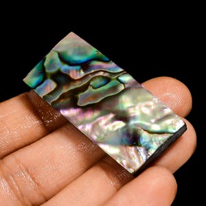 Può includere: Un pezzo rettangolare di conchiglia di abalone con colori iridescenti verdi, blu e viola. La superficie lucida riflette la luce, mostrando i motivi naturali. La conchiglia è tenuta su uno sfondo nero.