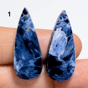 May include: Two teardrop-shaped gemstones with a polished surface. The stones are primarily dark blue with swirling patterns of lighter blue, black, and hints of gold. The number "1" is visible in the upper left corner.