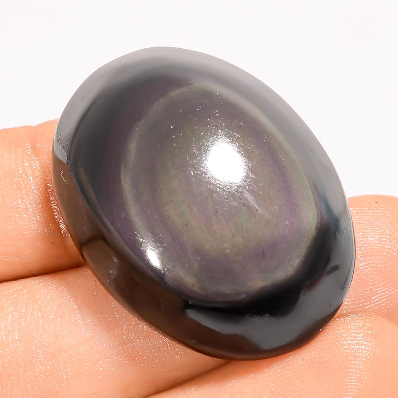 K&ouml;nnte beinhalten: Ein polierter, oval geformter Edelstein mit dunklem, schillerndem Glanz. Die Oberfl&auml;che des Steins reflektiert ein Farbspiel, einschlie&szlig;lich Lila und Gr&uuml;n, auf einem schwarzen Grund. Der Edelstein ist glatt und scheint in einer Hand gehalten zu werden.