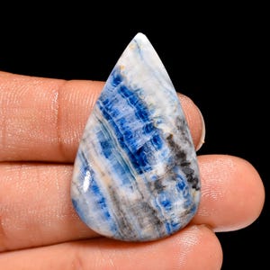 May include: A teardrop-shaped gemstone with a polished surface. The stone features a striking pattern of blue, white, gray, and black bands. The gemstone is held in a hand against a black background, highlighting its natural beauty.