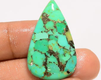 May include: A teardrop-shaped turquoise gemstone with a polished surface. The stone features a vibrant mix of light and dark turquoise hues, accented by veins of brown and gold. The gemstone is held in a hand.