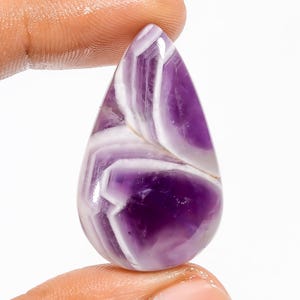 Puede incluir: Una gema pulida en forma de l&aacute;grima. La piedra presenta bandas de color blanco y p&uacute;rpura intenso, creando un patr&oacute;n rayado. La gema se sostiene entre dos dedos, mostrando su tama&ntilde;o y forma. Ideal para joyer&iacute;a.