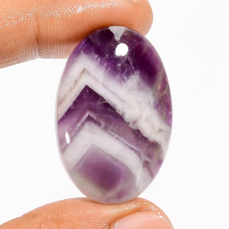 May include: An oval-shaped gemstone with a polished surface. The stone features a striking pattern of alternating bands of purple and white, creating a layered effect. The gemstone is held between two fingers.