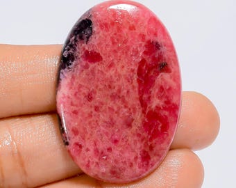 May include: Oval-shaped gemstone with a vibrant pink hue, accented by darker red and black inclusions. The polished surface reflects light, showcasing the stone's natural patterns and textures. The gemstone is held in a hand.