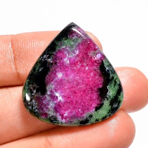 May include: A polished gemstone with a teardrop shape. The stone features a vibrant pink center, surrounded by green and black inclusions. The gemstone is held in a hand, showcasing its natural beauty and unique color variations.