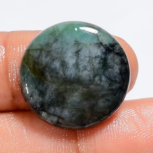 May include: A round, green emerald cabochon gemstone with black and white inclusions. The stone is polished and has a smooth, glossy surface.
