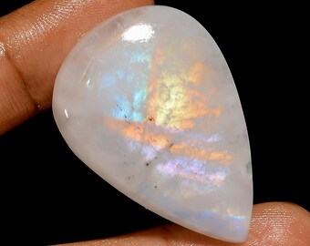 May include: A polished, teardrop-shaped gemstone with a milky white base. The stone displays iridescent flashes of orange, yellow, and blue light across its surface. The gem is held between two fingers against a black background.