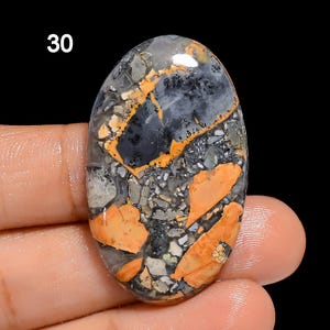 Puede incluir: Primer plano de una piedra ovalada colorida con una mezcla de colores negro, gris y naranja. La piedra tiene un patr&oacute;n moteado y est&aacute; engastada en un anillo.