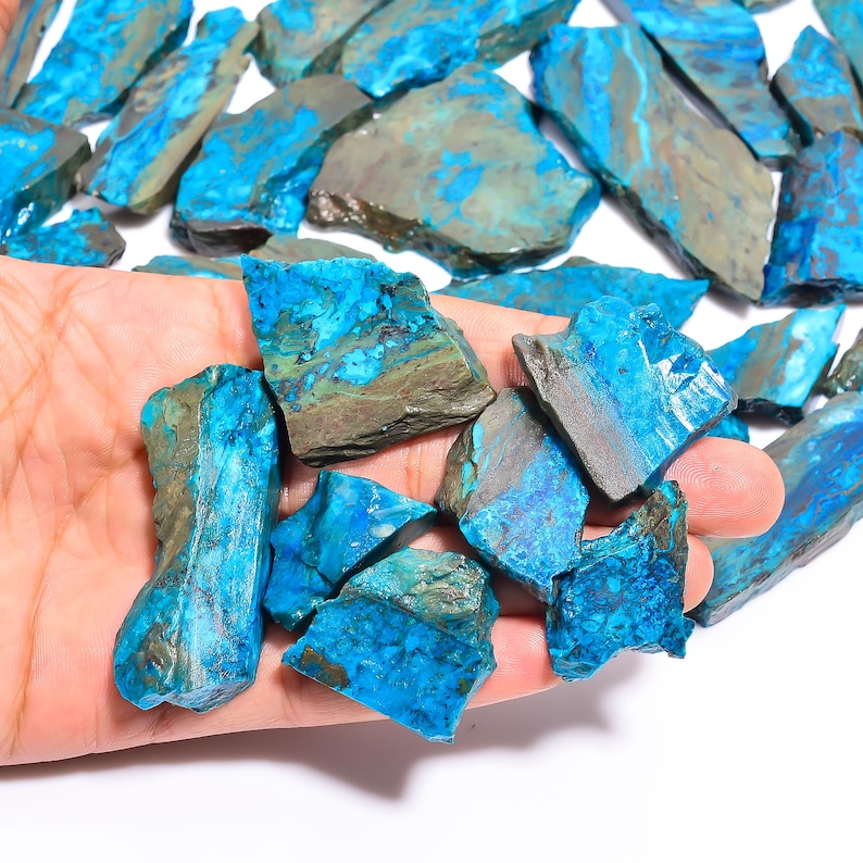 May include: A collection of raw, natural chrysocolla stones. The stones display a vibrant turquoise blue color with areas of brown and gray. The rough, irregular shapes of the stones are visible, showcasing their natural state. The stones are held in a hand.