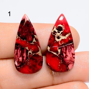 May include: Two teardrop-shaped red gemstones with dark red and black veining and metallic gold-colored inclusions. The stones are polished and smooth, featuring a vibrant crimson base color with intricate, branching patterns throughout.