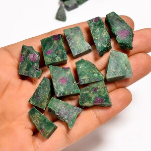 Może przedstawiać: Kolekcja surowo ciętych zielonych kamieni rubinu z widocznymi czerwonymi wrostkami. Kamienie różnią się rozmiarem i kształtem, z teksturowaną powierzchnią. Kamienie szlachetne są trzymane w dłoni, ukazując ich naturalne piękno.