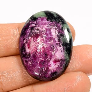 May include: An oval-shaped gemstone with a polished surface. The stone features a blend of colors, including deep purple, black, and green. The purple area has a crystalline appearance, with white and pink inclusions. The gemstone is held in a hand.