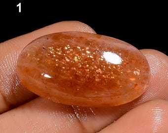 May include: An oval-shaped sunstone gemstone with a smooth, polished cabochon surface. The stone displays a translucent orange hue with internal metallic copper-colored inclusions that create a sparkling aventurescence effect under light.