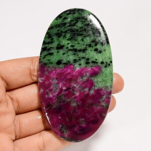 May include: An oval-shaped gemstone with a polished surface. The top half is green with black speckles, and the bottom half is a vibrant magenta. The stone is held in a hand, showcasing its natural beauty and unique color combination.