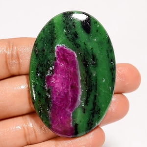 May include: Oval-shaped ruby zoisite gemstone. The polished stone features a vibrant green base with black inclusions and a prominent magenta ruby center. The gemstone is held in a hand, showcasing its natural beauty and color variations.