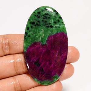 May include: An oval-shaped gemstone featuring a vibrant color palette. The top half is a rich green speckled with black, transitioning to a deep magenta at the bottom. The polished surface reflects light, highlighting the stone's natural beauty.