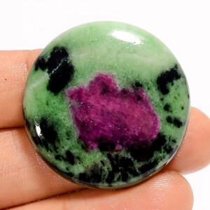 May include: A polished, round gemstone featuring a vibrant mix of colors. The stone has a light green base with dark black spots and a central patch of deep magenta. The gemstone is held in a hand.