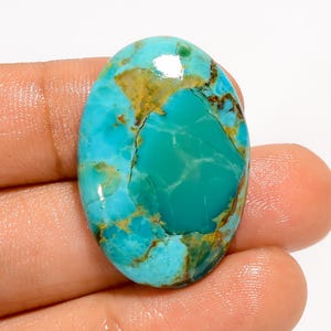 May include: Oval-shaped turquoise gemstone with a polished surface. The stone features a mix of vibrant blue and green hues, with brown and gold veining throughout. The gemstone is held in a hand, showcasing its natural beauty and unique patterns.