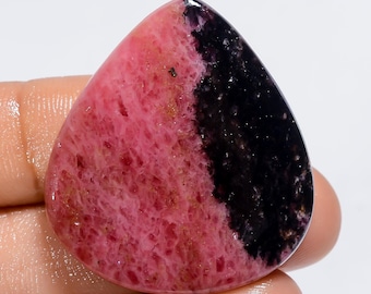 Könnte beinhalten: Ein rosa-schwarzer Cabochon-Edelstein in Tropfenform. Der Edelstein hat eine raue, strukturierte Oberfläche.