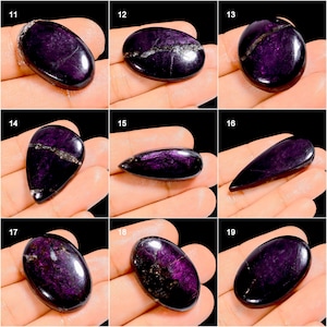 May include: A collection of polished, oval and teardrop-shaped gemstones in varying sizes. The stones are a deep purple color with streaks of silver and white. The gemstones are displayed on a hand, showcasing their smooth, reflective surfaces.