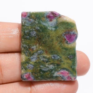 May include: A rectangular gemstone with a rough, natural edge. The stone features a mottled pattern of green, blue, and yellow hues, with vibrant red ruby inclusions scattered throughout. The stone is held in a hand.