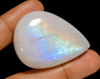 May include: A polished, teardrop-shaped gemstone with a pearly white base and iridescent flashes of blue, yellow, and orange. The stone has a smooth, glossy surface and is held against a black background.