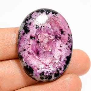 May include: Oval-shaped gemstone with a polished surface. The stone features a blend of pink and black colors, with a speckled pattern. The pink areas have a crystalline appearance, while the black sections are more opaque. The gemstone is held in a hand.