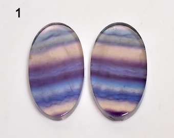 Puede incluir: Dos piedras preciosas ovaladas de fluorita con capas de tonos morados, azules y crema. Las piedras pulidas tienen un acabado liso y brillante. El número "1" es visible en la esquina superior izquierda. Ideal para la fabricación de joyas.