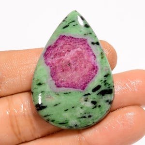 May include: A polished, teardrop-shaped gemstone featuring a vibrant pink center surrounded by a green matrix with black inclusions. The stone's surface is smooth and reflective, showcasing a natural, organic pattern. The gemstone is held in a hand.