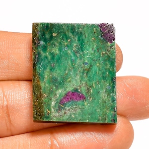 May include: A rectangular gemstone with a vibrant green base color, speckled with small blue and brown inclusions. Deep magenta-colored patches are visible within the stone. The gemstone is held in a hand.