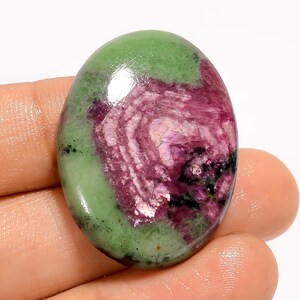 May include: Oval-shaped gemstone with a polished surface. The stone features a blend of colors, including a vibrant green, deep magenta, and hints of black. The magenta section has a swirling pattern. The gemstone is held in a hand.