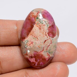 Op de afbeelding: Een ovaalvormige edelsteen met een mix van kleuren. De steen heeft een mix van roze, rood, beige en groene tinten, met een gebarsten, getextureerd uiterlijk. De edelsteen wordt in een hand gehouden.