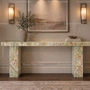 Puede incluir: Una consola rectangular con una superficie de color verde claro y blanco similar al mármol. La mesa tiene dos patas rectangulares y está colocada en un pasillo bien iluminado. Un jarrón con vegetación y una pintura están en la pared.