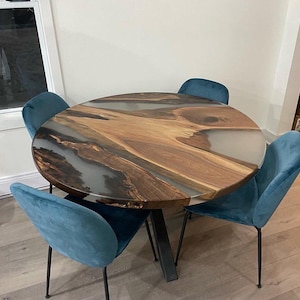 Puede incluir: Una mesa redonda de comedor de madera con una tapa de resina epoxi transparente. La mesa tiene un patrón de veta de madera natural y está rodeada por cuatro sillas de terciopelo azul turquesa con patas de metal negro.