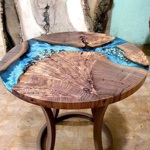 Puede incluir: Mesa auxiliar redonda de madera con un diseño único. La tapa presenta un río de resina azul que fluye a través de la veta natural de la madera. La mesa tiene una base curva de color marrón oscuro. La mesa tiene aproximadamente 61 cm de diámetro.