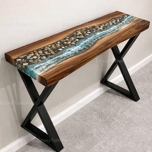 Può includere: Una consolle in legno con un design a fiume in resina epossidica. Il tavolo presenta un piano in legno scuro con un fiume centrale di resina blu-verde e pietre incastonate. Le gambe in metallo nero a forma di X sostengono il tavolo.