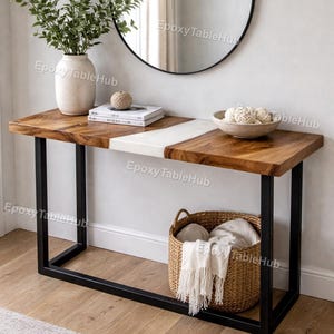 Puede incluir: Una consola de madera con una franja de epoxi blanca, sostenida por patas de metal negro. La mesa está decorada con un jarrón de vegetación, libros y un cuenco con esferas decorativas. Debajo hay una cesta de mimbre con una manta.