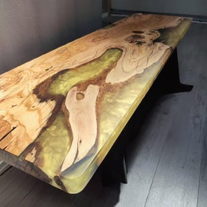 Op de afbeelding: Handgemaakte houten tafel met een uniek ontwerp. Het tafelblad heeft natuurlijke houtnerfpatronen en een rivier van doorschijnende geelgroene hars. De tafel wordt ondersteund door zwarte poten, wat een moderne esthetiek creëert.