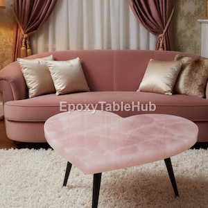 Puede incluir: Una mesa de centro rosa en forma de corazón con patas negras se encuentra sobre una alfombra blanca. Un sofá rosa con cojines dorados está detrás de la mesa. Una lámpara, flores y una chimenea completan la decoración de la habitación.