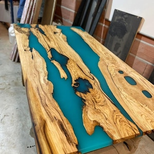 Op de afbeelding: Een houten tafelblad met een inleg van turquoiseblauwe epoxyhars. De hars stroomt in een rivierachtig patroon door het hout.